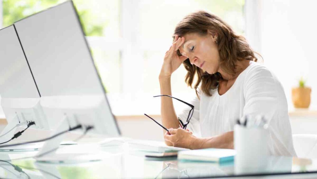Woman wearing white sitting at desk in front of a computer holding her head looking stressed from being behind on bookkeeping.
