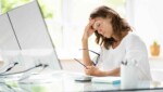 Woman wearing white sitting at desk in front of a computer holding her head looking stressed from being behind on bookkeeping.