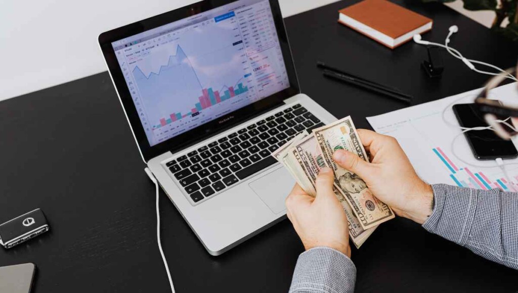 Person counting cash in hand at a desk with a laptop displaying stock market charts and financial data in the background, checking their profit vs. cash flow.